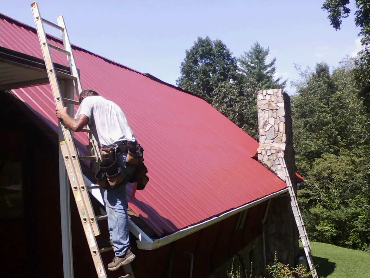 Roofer on a ladder beside a red metal roof and stone chimney — Soffit & Fascia Repair by Roofing Brothers in White Swan